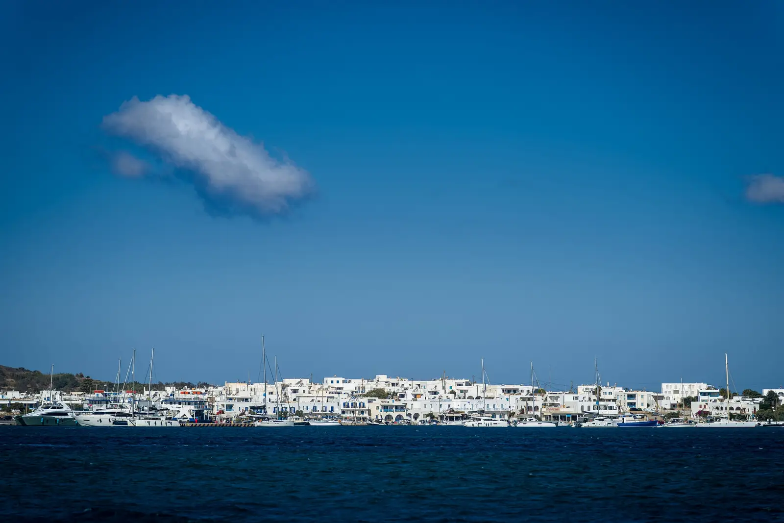 A white harbour town with sailing boats seen across deep blue water, one small cloud in the sky