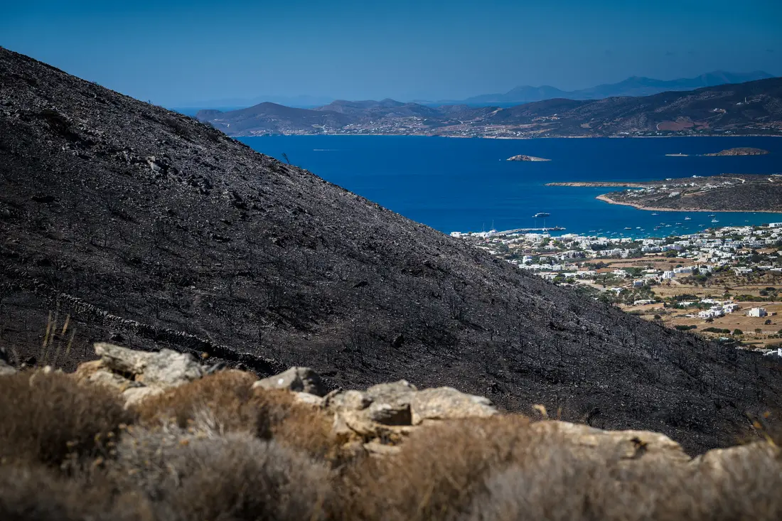 A burnt black hillside dropping towards a blue bay, a white coastal town and islands beyond