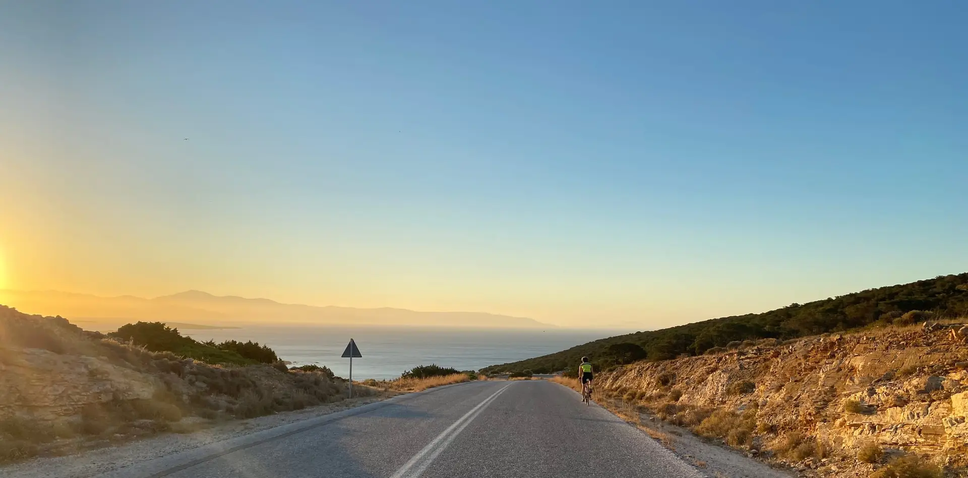 An empty road running down to the sea in low sunlight, a lone cyclist in the distance