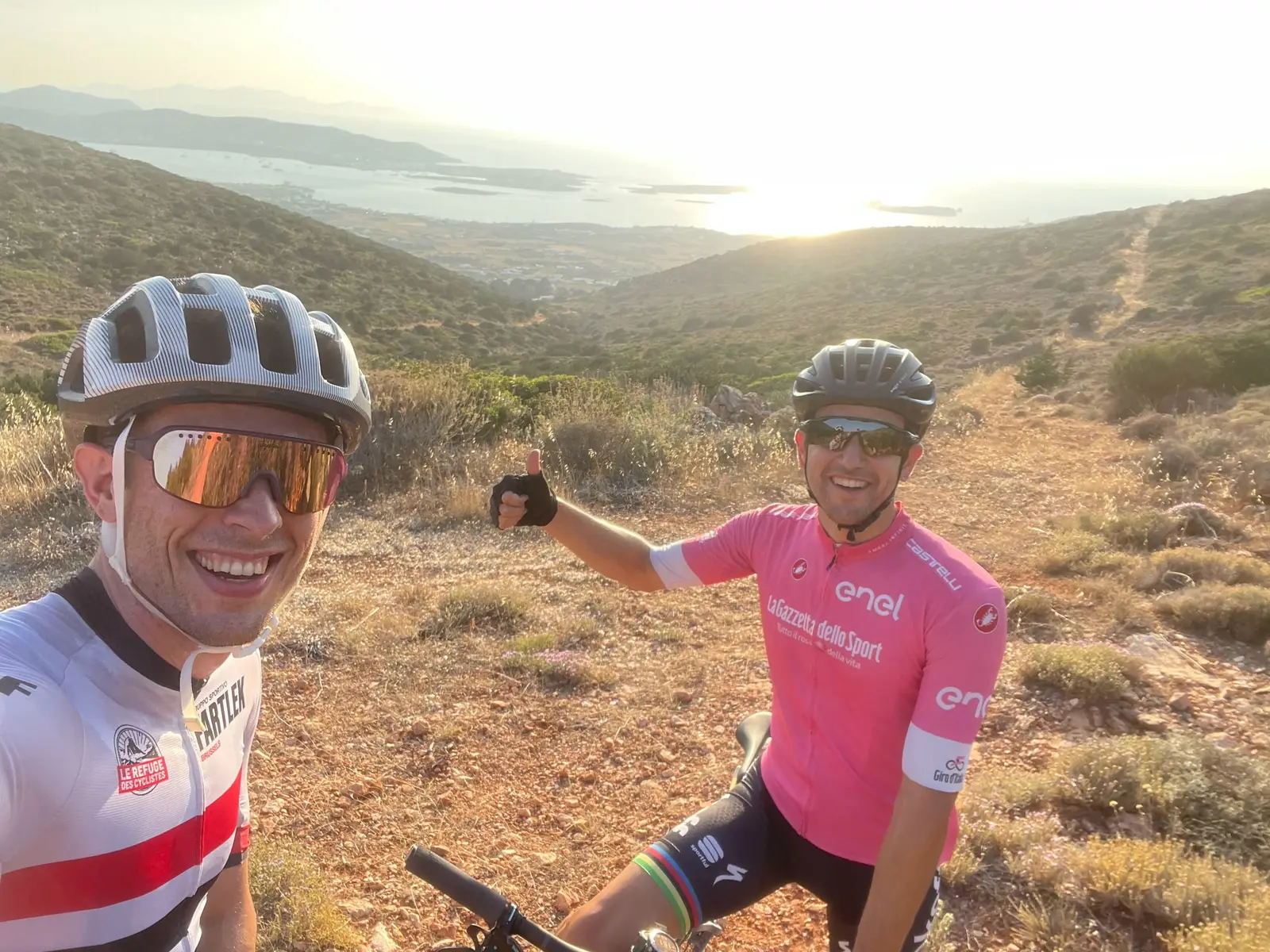 Sam and Panos in cycling kit taking a selfie on a hilltop above the sea